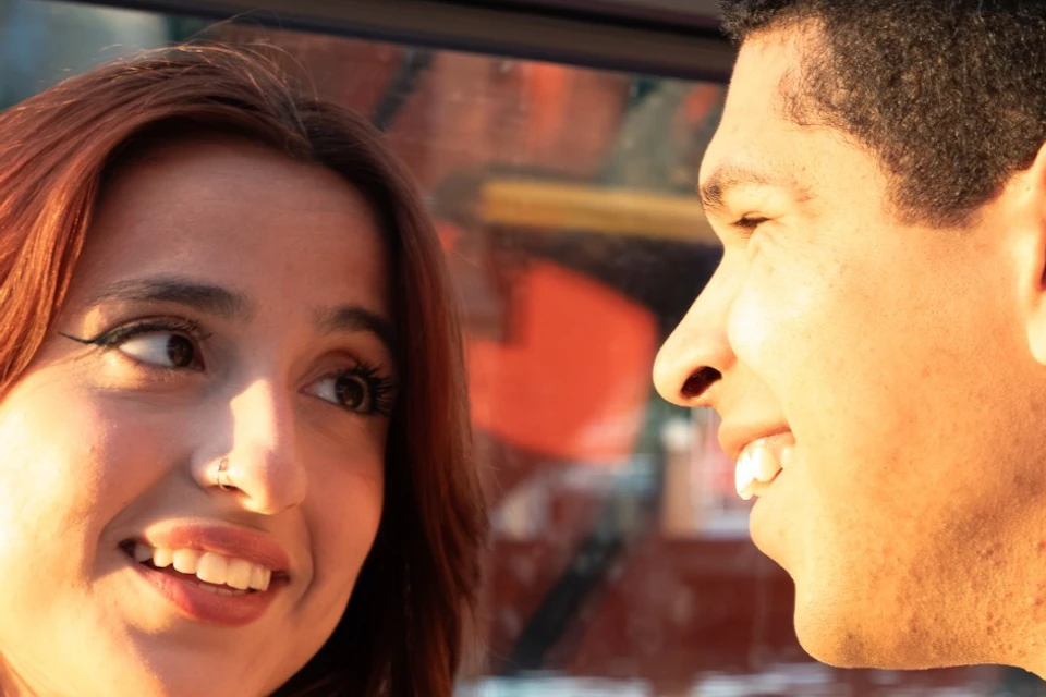 Two people smiling at each other while holding wine glasses, seated inside a boat with a colorful ship visible outside.