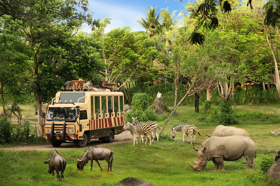A safari truck drives through a lush landscape with zebras, rhinoceroses, and wildebeests grazing around.