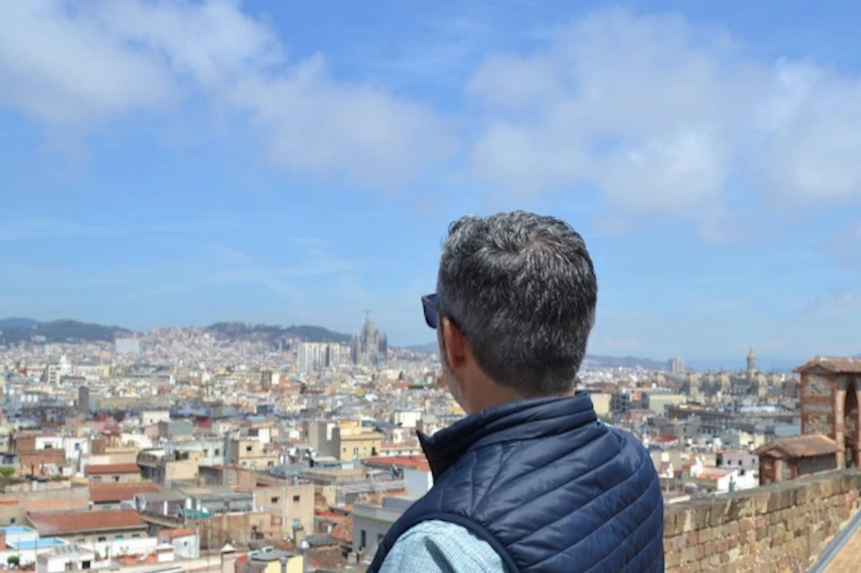 A person wearing a blue vest and sunglasses looks out over a cityscape under a partly cloudy sky.