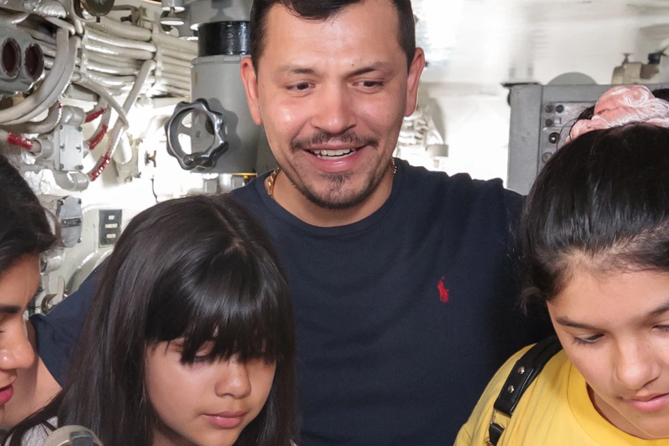 A man and three children inside a submarine, interacting with equipment and controls in a confined technical space.