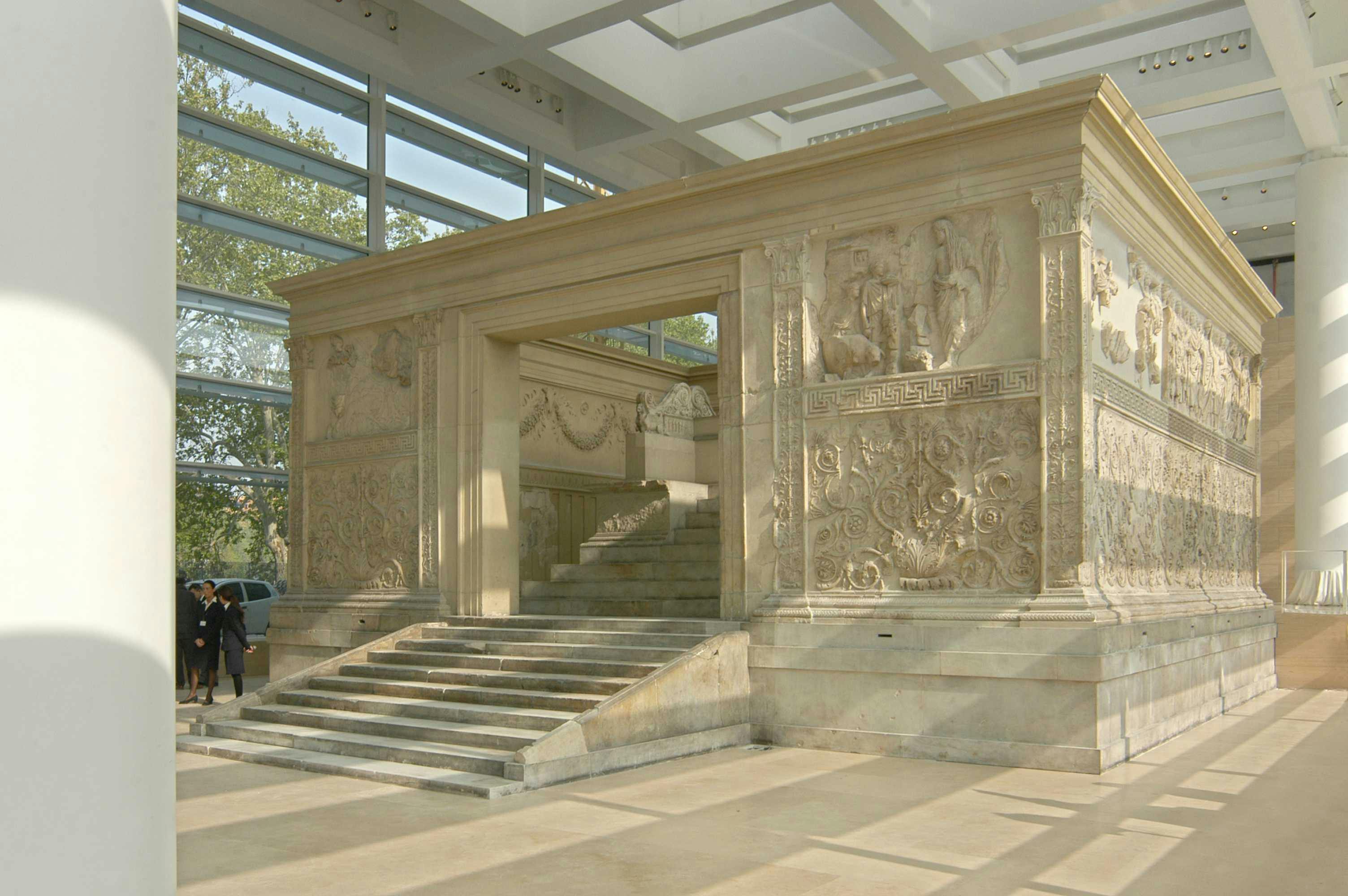 Ancient stone structure indoors, featuring carved reliefs, steps, and a large doorway. Natural light from large windows.
