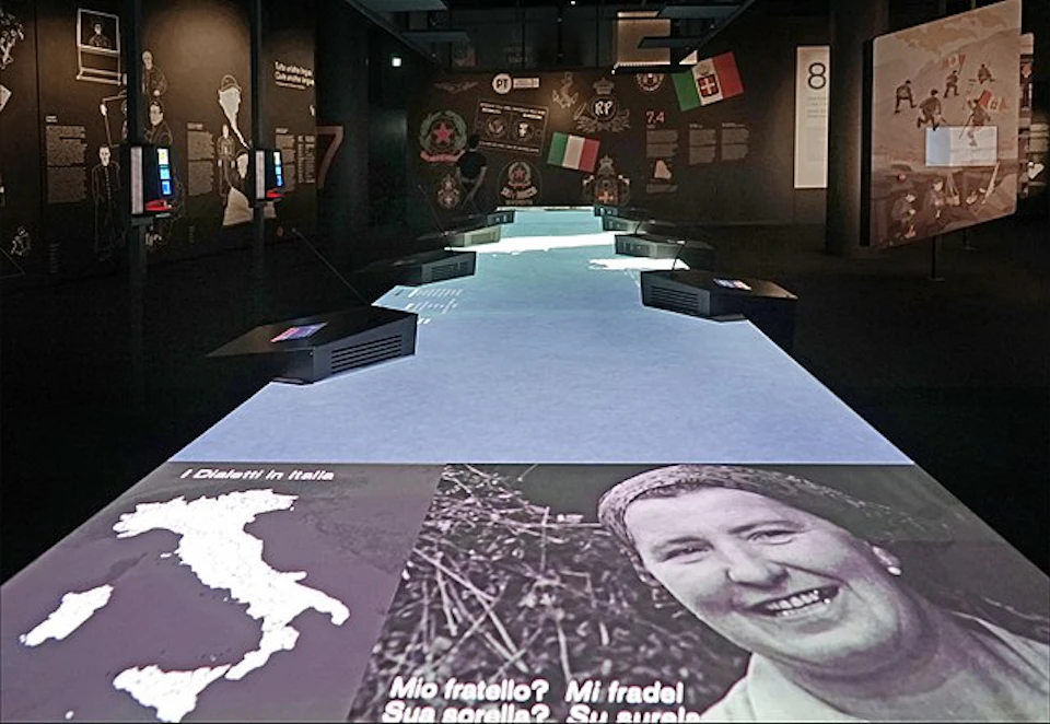 Interactive exhibit featuring historical imagery, a map of Italy, and text in Italian. Flag displays and projection screens present.