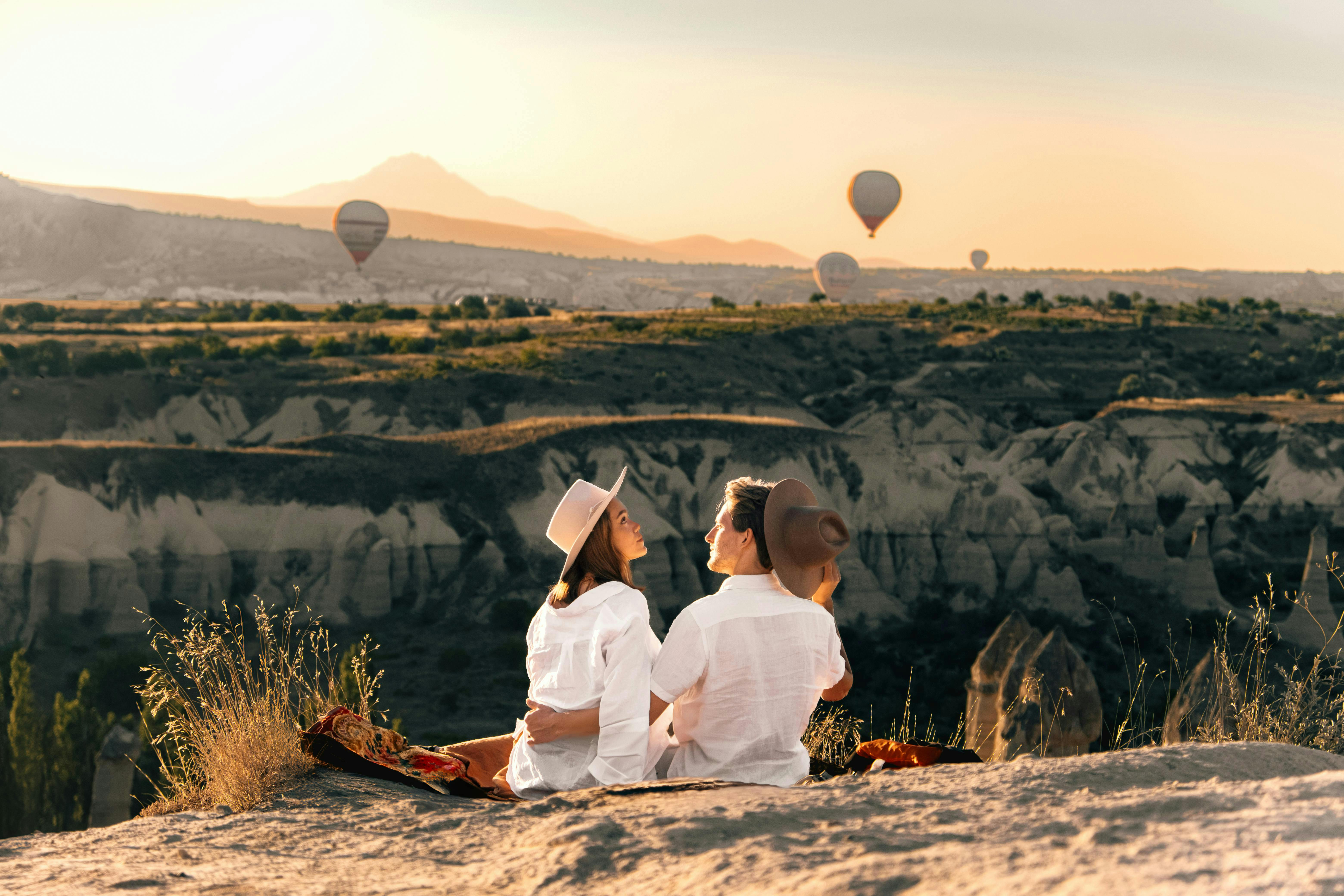Un couple en chemise blanche et chapeau est assis sur une falaise surplombant un paysage de montgolfières au coucher du soleil.