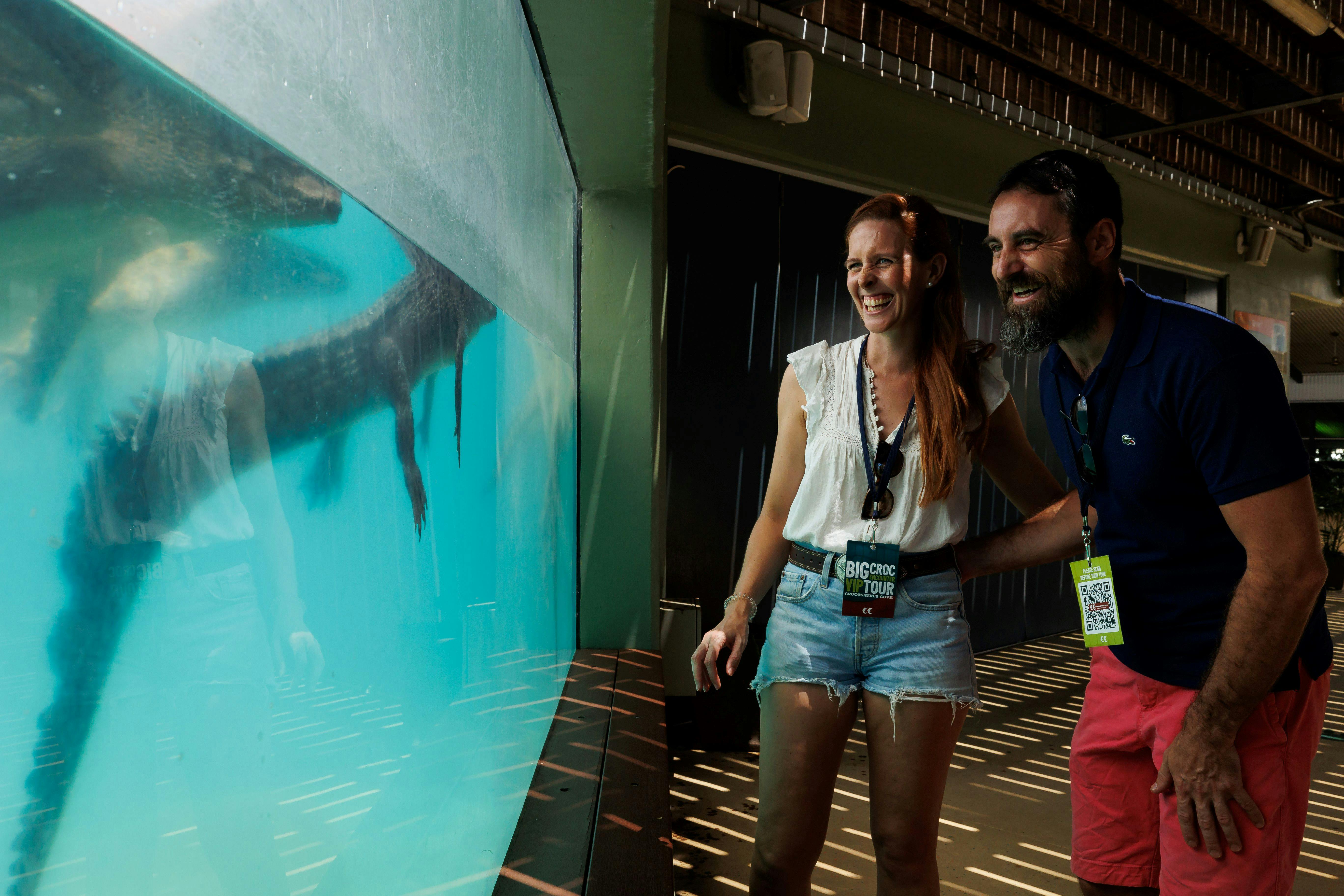 Två personer ler och tittar genom ett stort akvariefönster, med en som bär en &#34;Big Croc Tour&#34; -snodd.