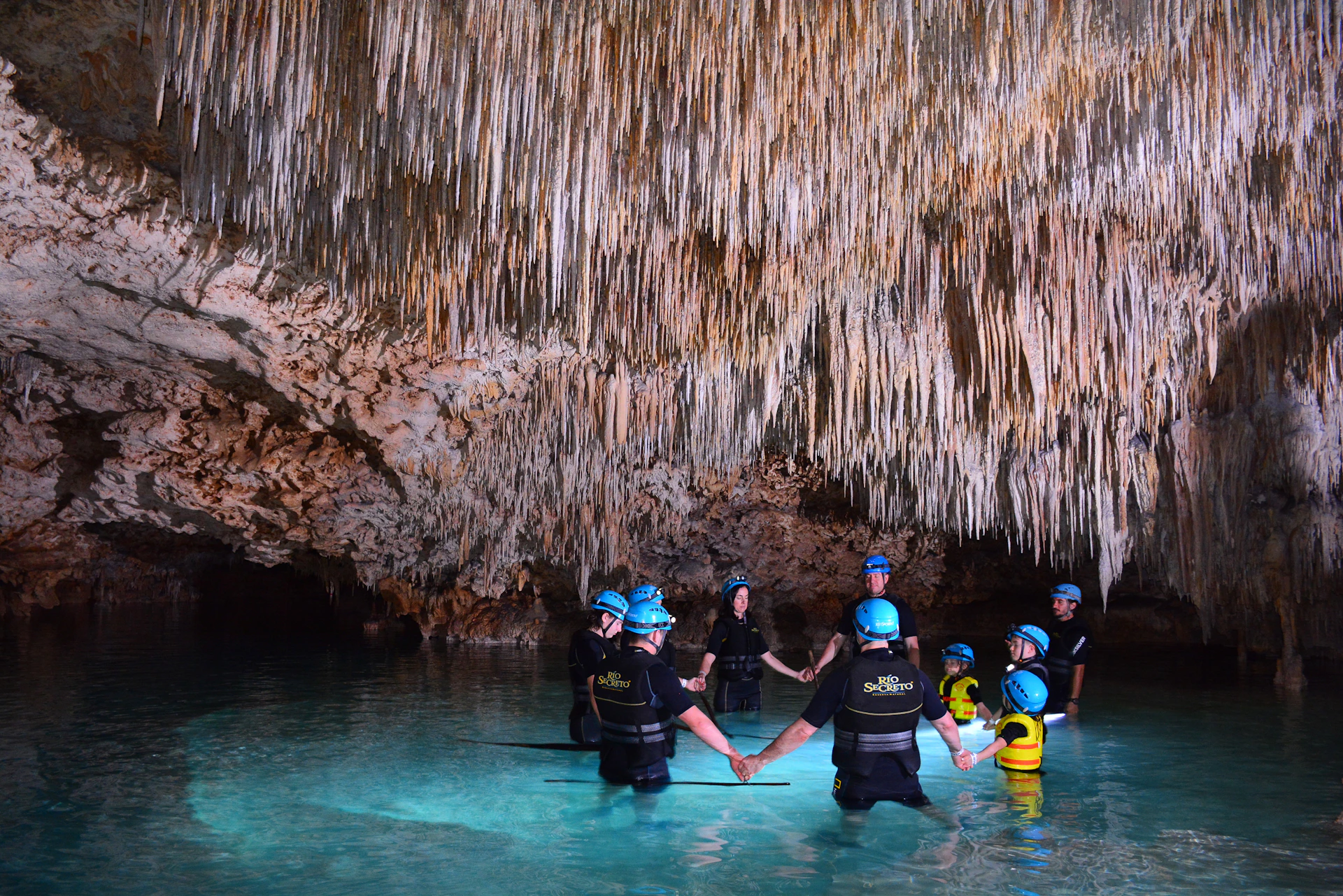 A group of people in helmets and protective gear hold hands while standing in shallow, clear water inside a cave with stalactites.