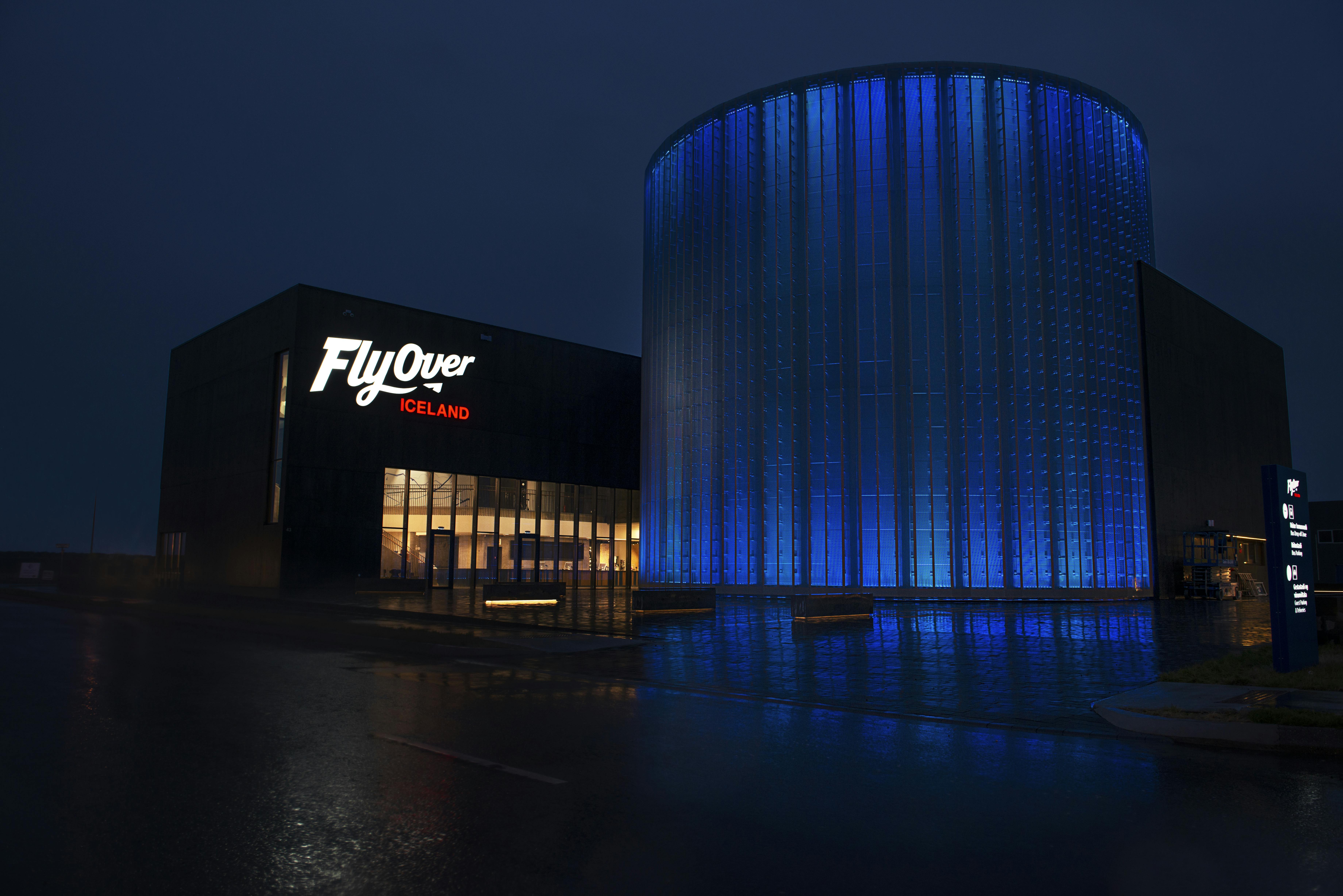 Η είσοδος του FlyOver Iceland τη νύχτα, με μια μεγάλη φωτεινή πινακίδα και ένα κυλινδρικό κτίριο φωτισμένο με μπλε χρώμα.