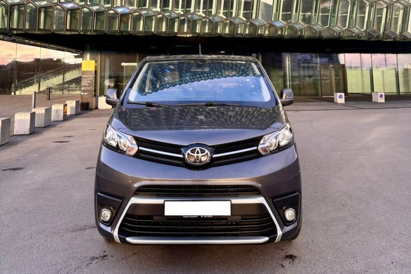 Front view of a grey Toyota minivan parked on a street with a modern glass building in the background.