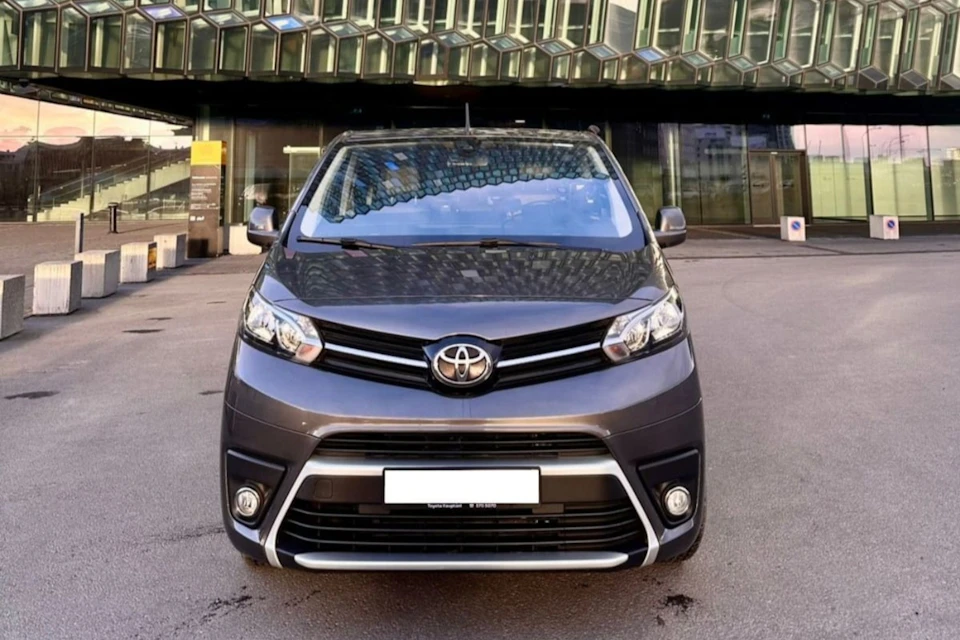 Front view of a grey Toyota minivan parked on a street with a modern glass building in the background.