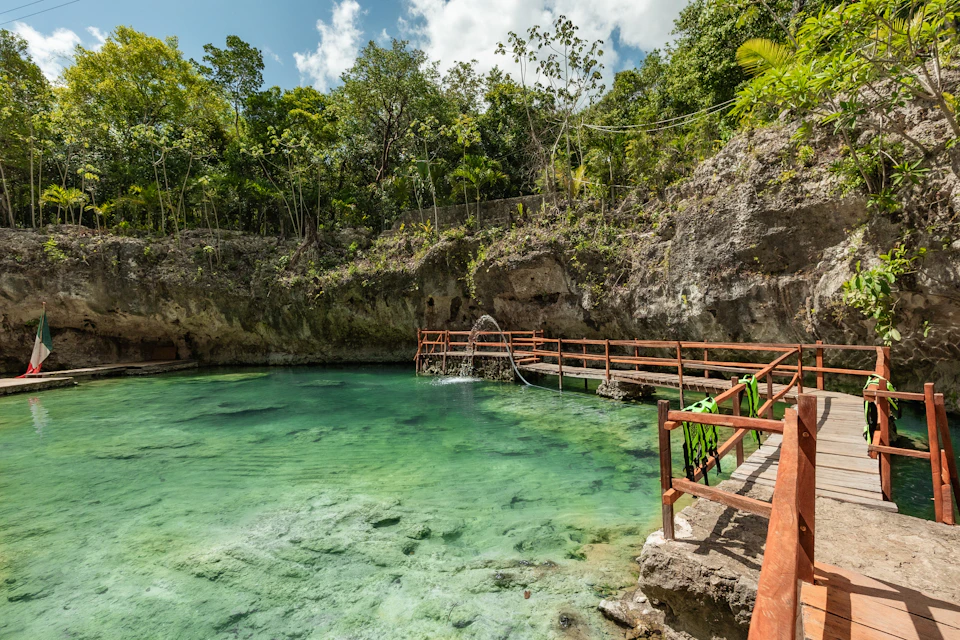 A clear natural pool surrounded by rocky cliffs and lush greenery, with a wooden walkway and steps leading into the water.