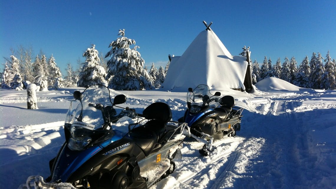 Δύο οχήματα χιονιού σταθμευμένα μπροστά από μια χιονισμένη σκηνή σε ένα χιονισμένο τοπίο με χιονισμένα δέντρα κάτω από έναν καταγάλανο ουρανό.