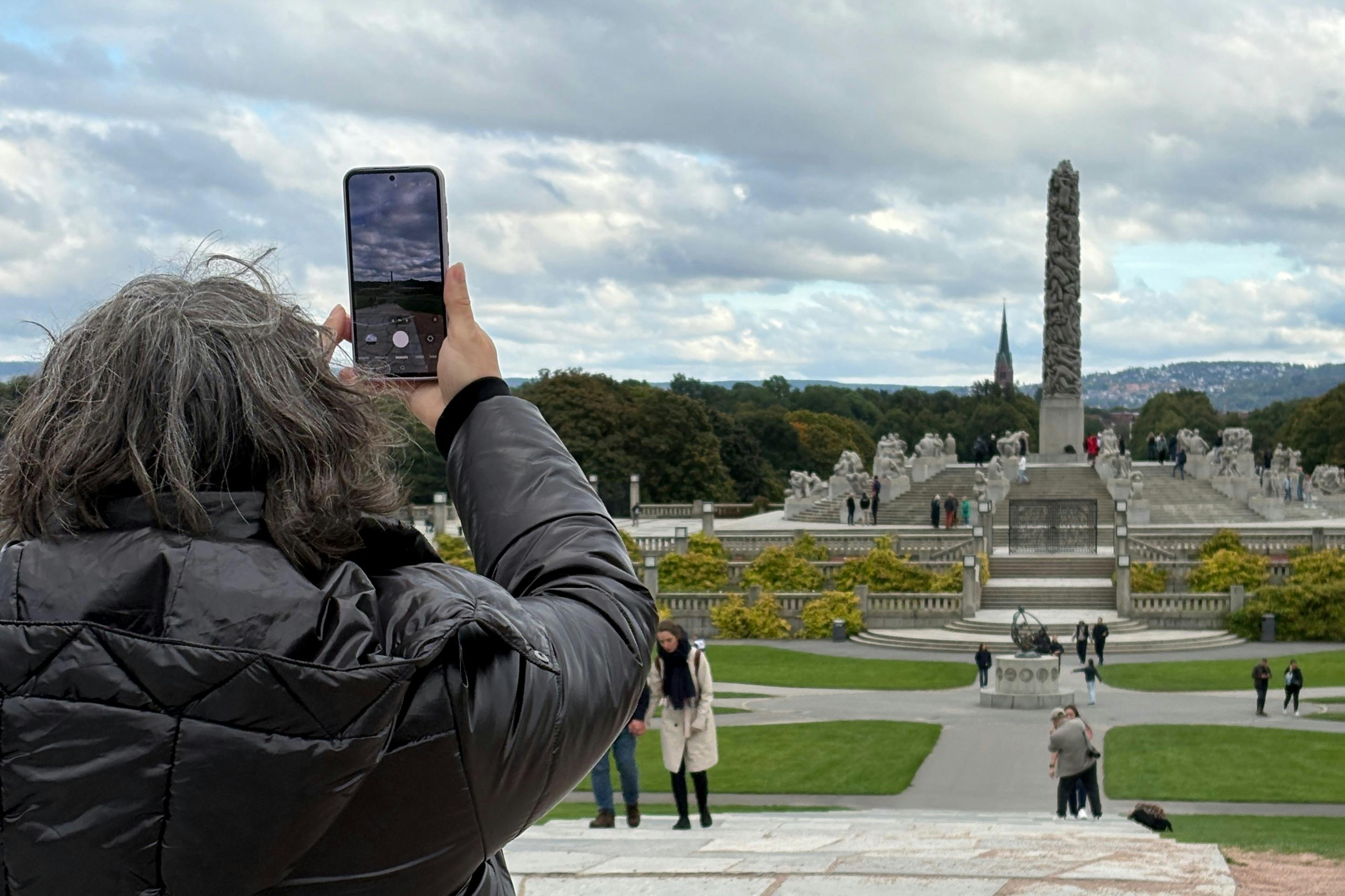 穿着黑色外套的人在多云的天空下拍摄一个公园，里面有雕像、绿色植物和方尖碑。