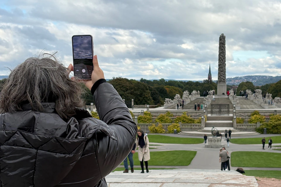 Person in black coat photographing a park with statues, greenery, and an obelisk under a cloudy sky.