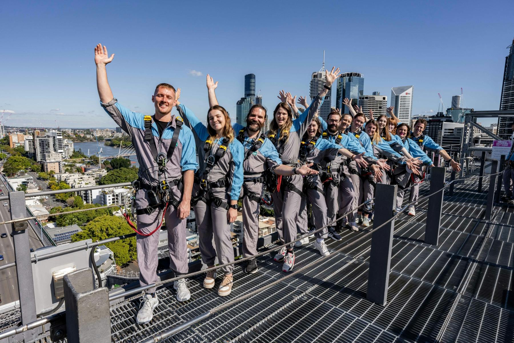 一群戴着安全带的人在一个高楼大厦的城市屋顶上挥手，背景是一条河流。