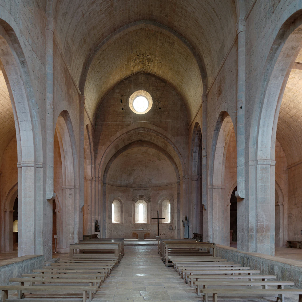Interior of a stone church with vaulted ceilings, wooden benches, arches, a round window, and a cross at the altar.