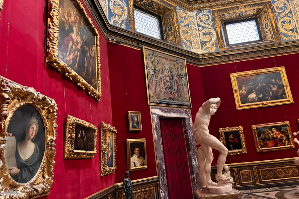 A museum gallery with red walls, golden-framed paintings, ornate ceiling, and a classical marble statue in the foreground.