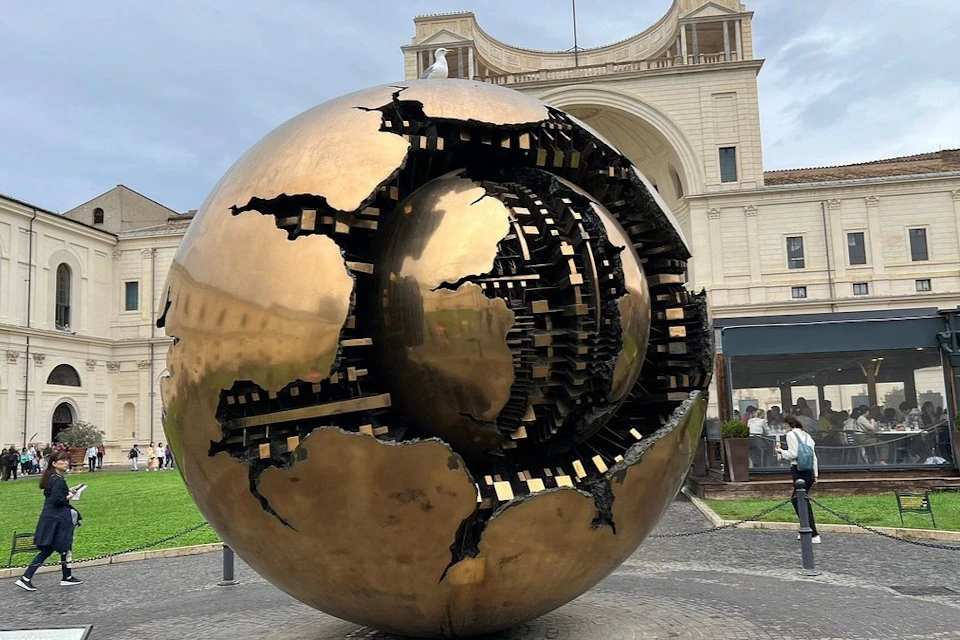 A large bronze spherical sculpture with intricate inner details, set in a plaza near a classical-style building.