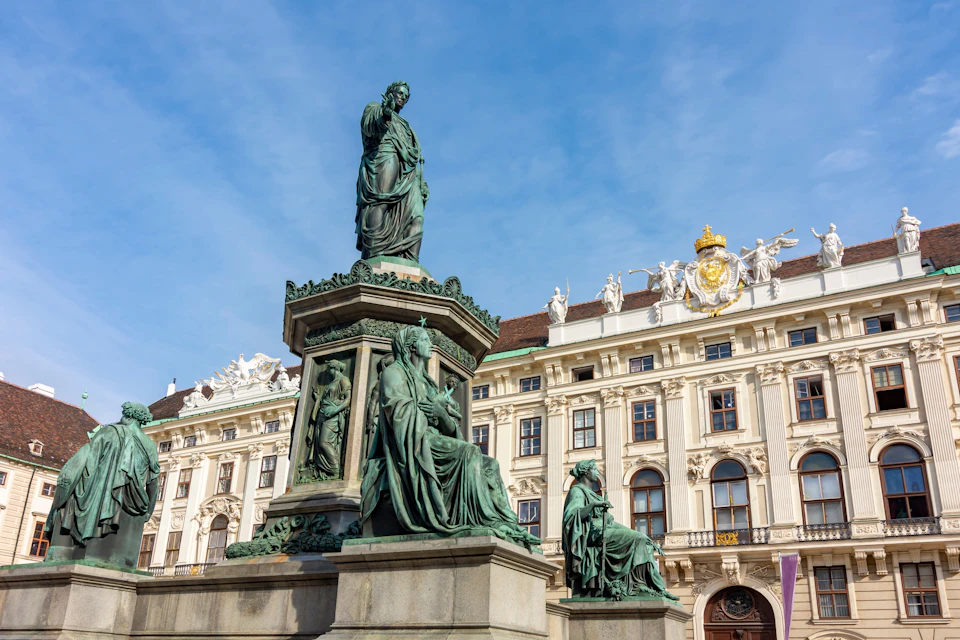 Bronze statues on a pedestal in front of an ornate building with sculptures and a golden crown under a blue sky.