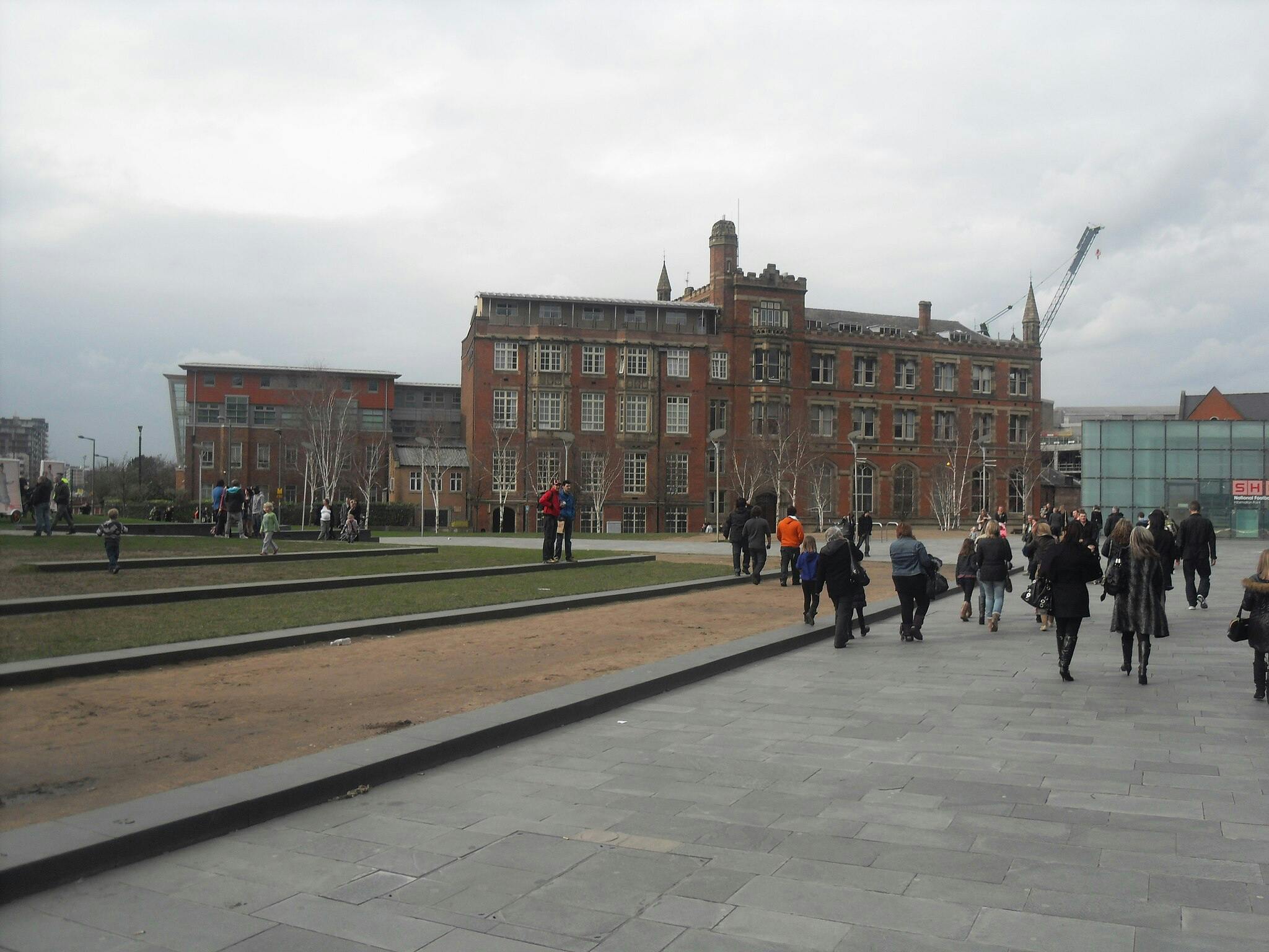 Folk, der går og hygger sig i en park med asfalterede stier og en rød murstensbygning i baggrunden på en overskyet dag.
