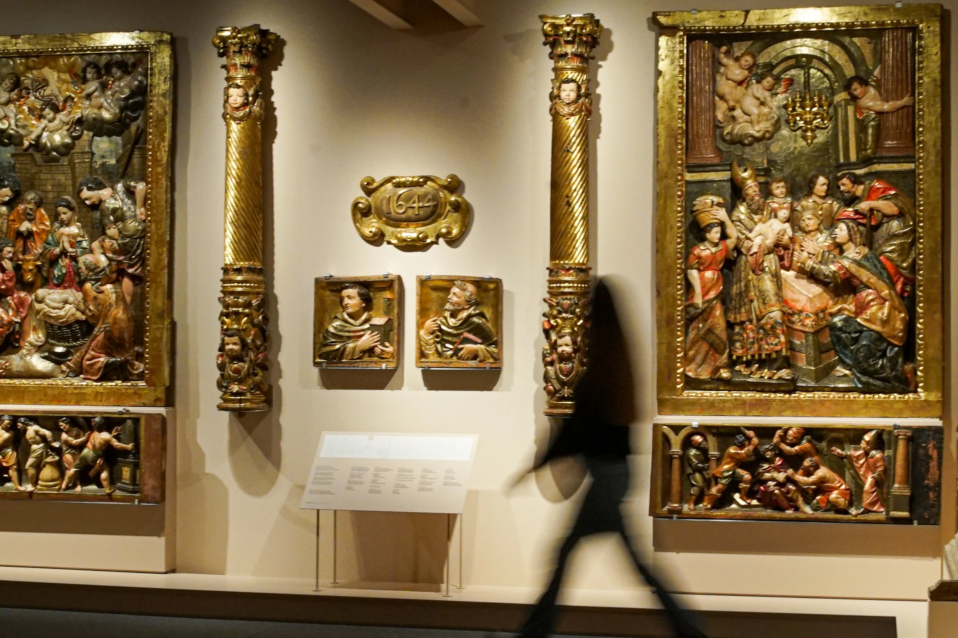 A blurred figure walks past a wall display of ornate, gold-framed religious artwork and sculptures in a museum.