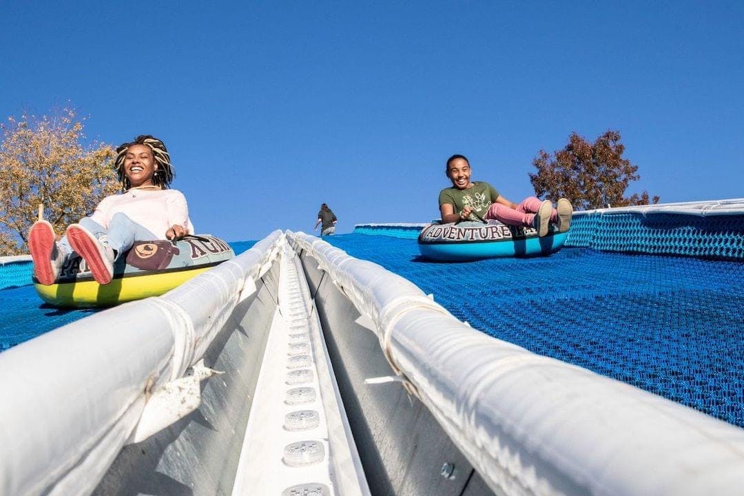 Two people slide down an inclined track on inflatable tubes, smiling. A tree and a clear blue sky are visible in the background.