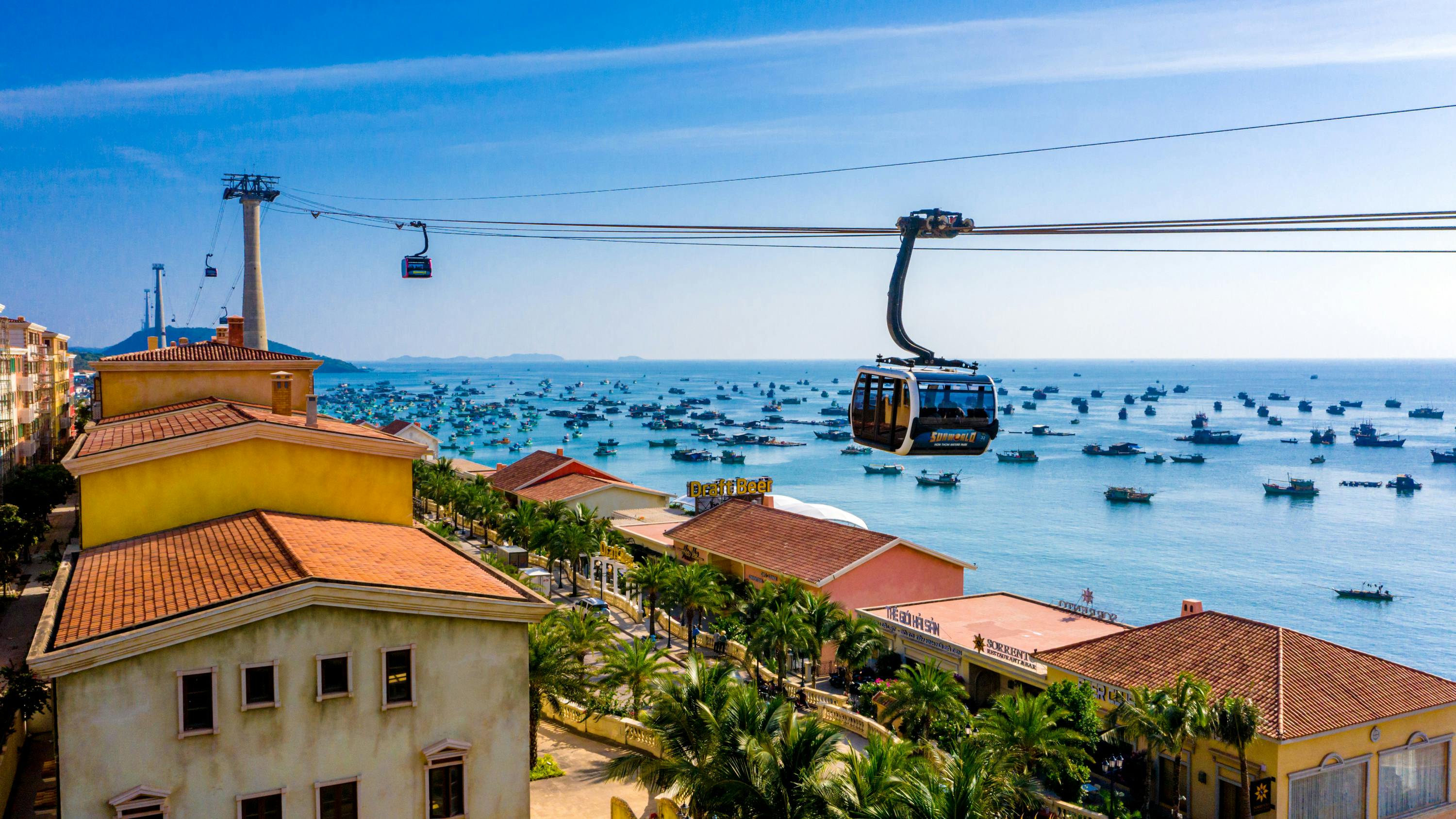 A cable car travels above rooftops and palm trees, overlooking a sea filled with numerous boats under a clear blue sky.