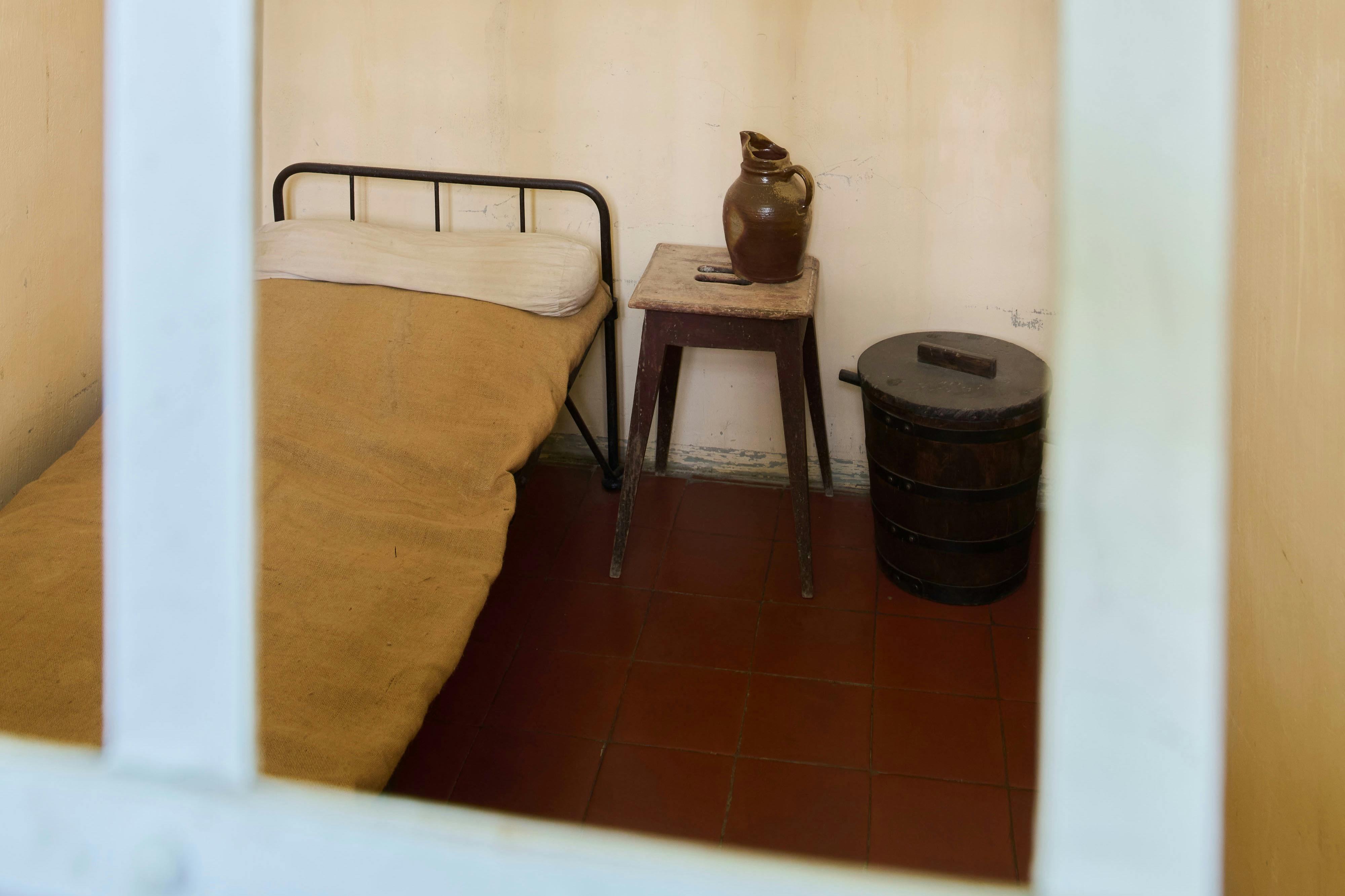 简单的房间，配有金属框架床、黄色毯子、带粘土壶的木凳和瓷砖地板上的深色木桶。