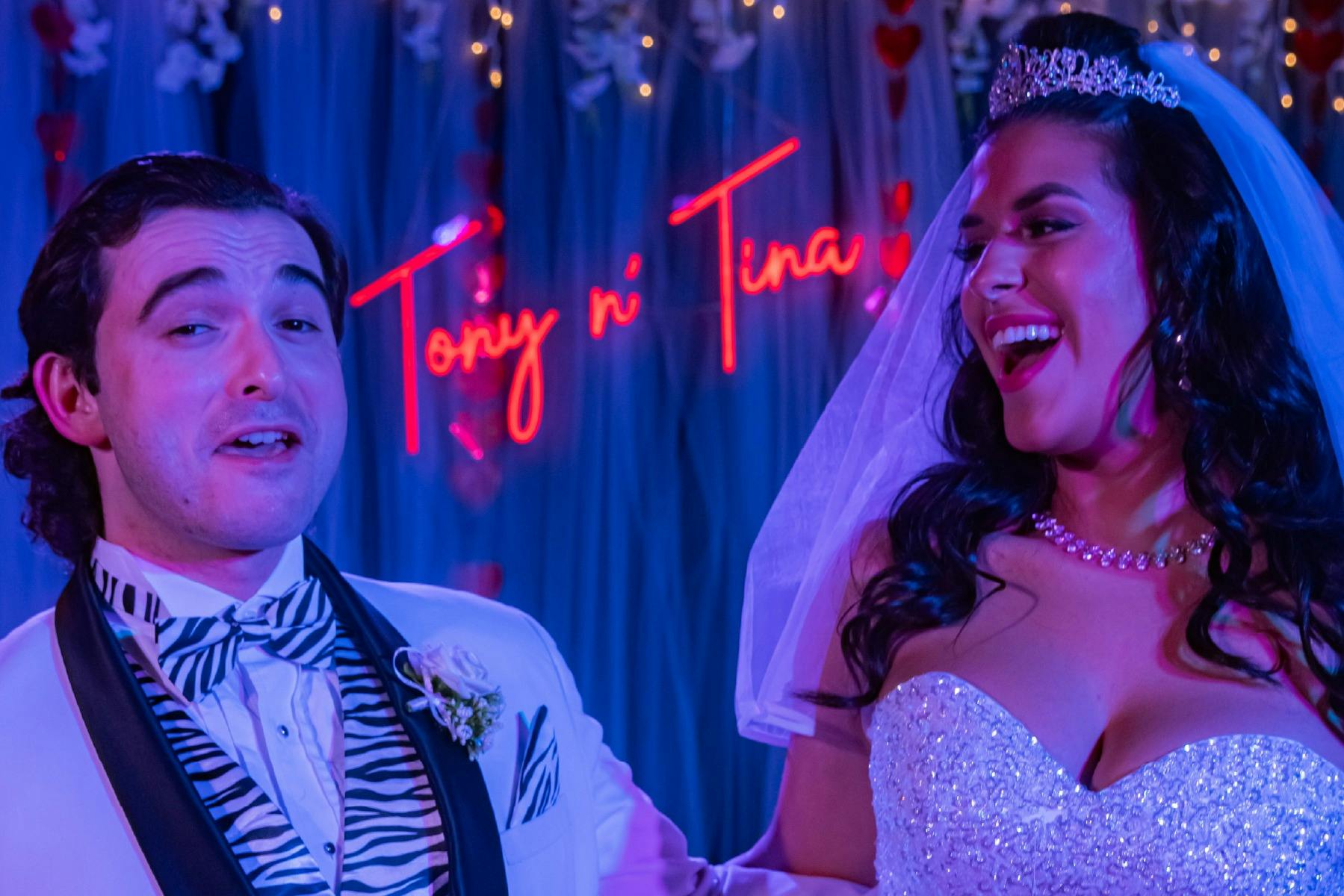 A bride and groom smiling in front of a neon sign that reads "Tony n' Tina," with blue and red lighting in the background.