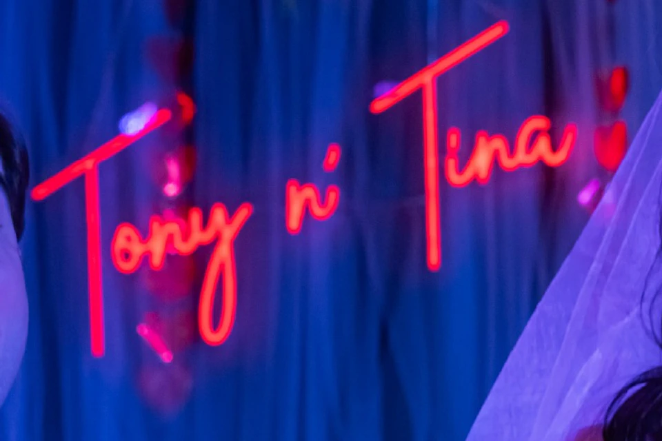 A bride and groom smiling in front of a neon sign that reads "Tony n' Tina," with blue and red lighting in the background.
