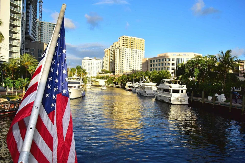 A waterway lined with yachts and buildings under a blue sky, with an American flag in the foreground.