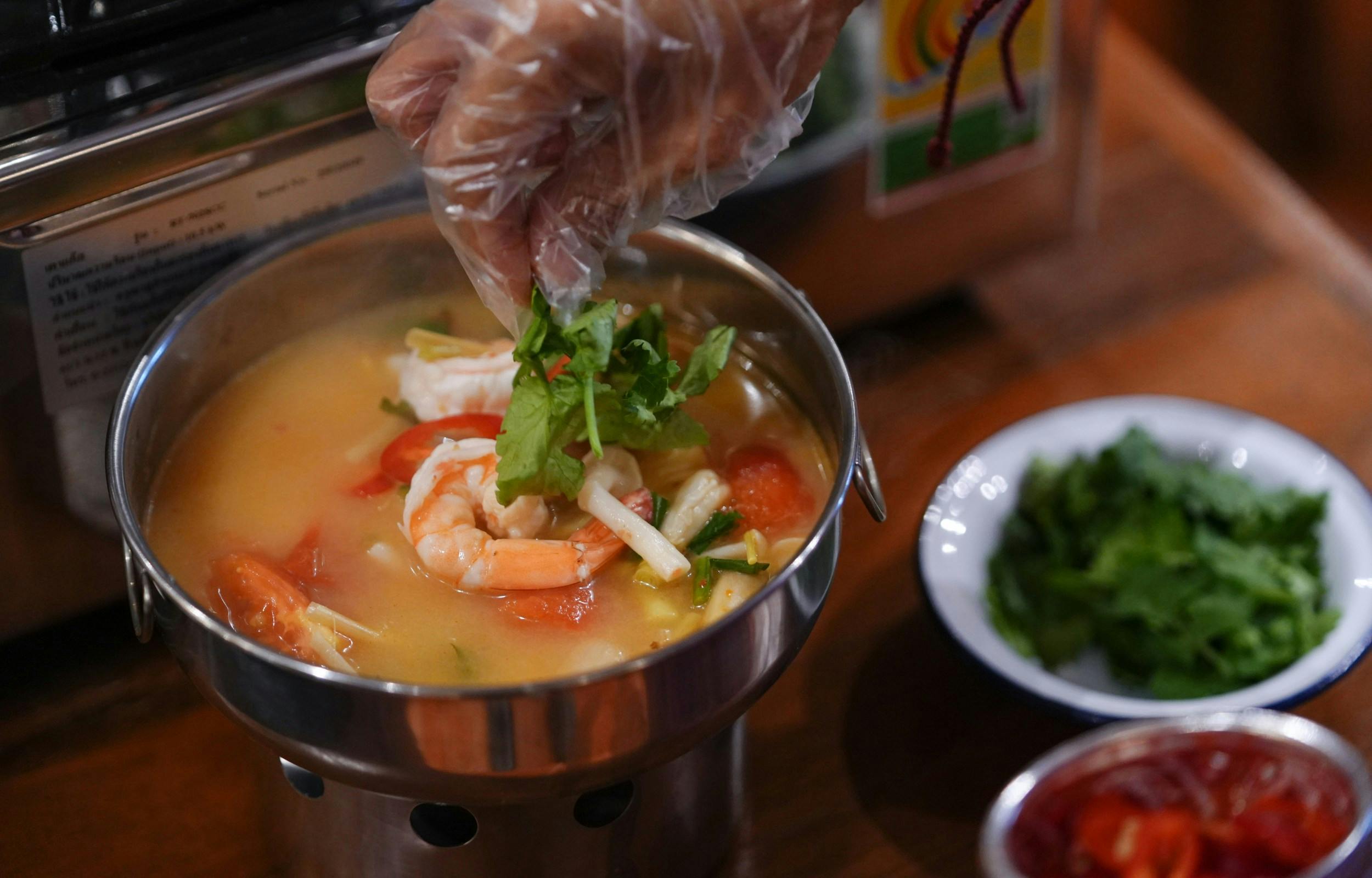 A steaming bowl of soup with shrimp, vegetables, and herbs being garnished by a hand wearing a transparent glove.