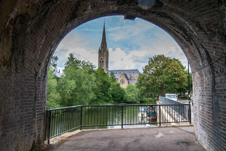 View through a brick archway of a church with a tall spire, trees, and a river with a boat and a railing in the foreground.