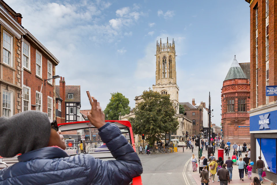 Person taking a photo of a church tower from the top of a red double-decker bus in a busy street with pedestrians.