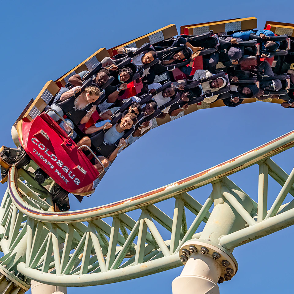 People riding a Colossus roller coaster, upside down on a loop, with expressions of excitement under a clear blue sky.