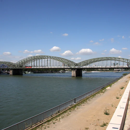 En grön järnbro sträcker sig över en bred flod, med en tågövergång. Människor går längs en flodväg under en klar, blå himmel.
