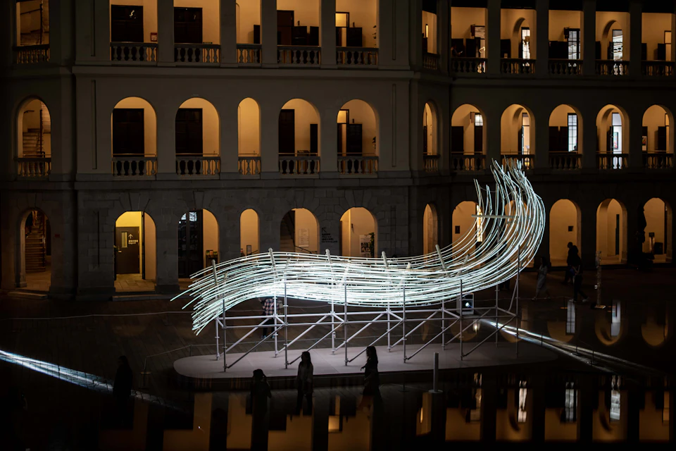 An illuminated modern art installation resembling a flowing wave, set in the courtyard of a building with arched windows.