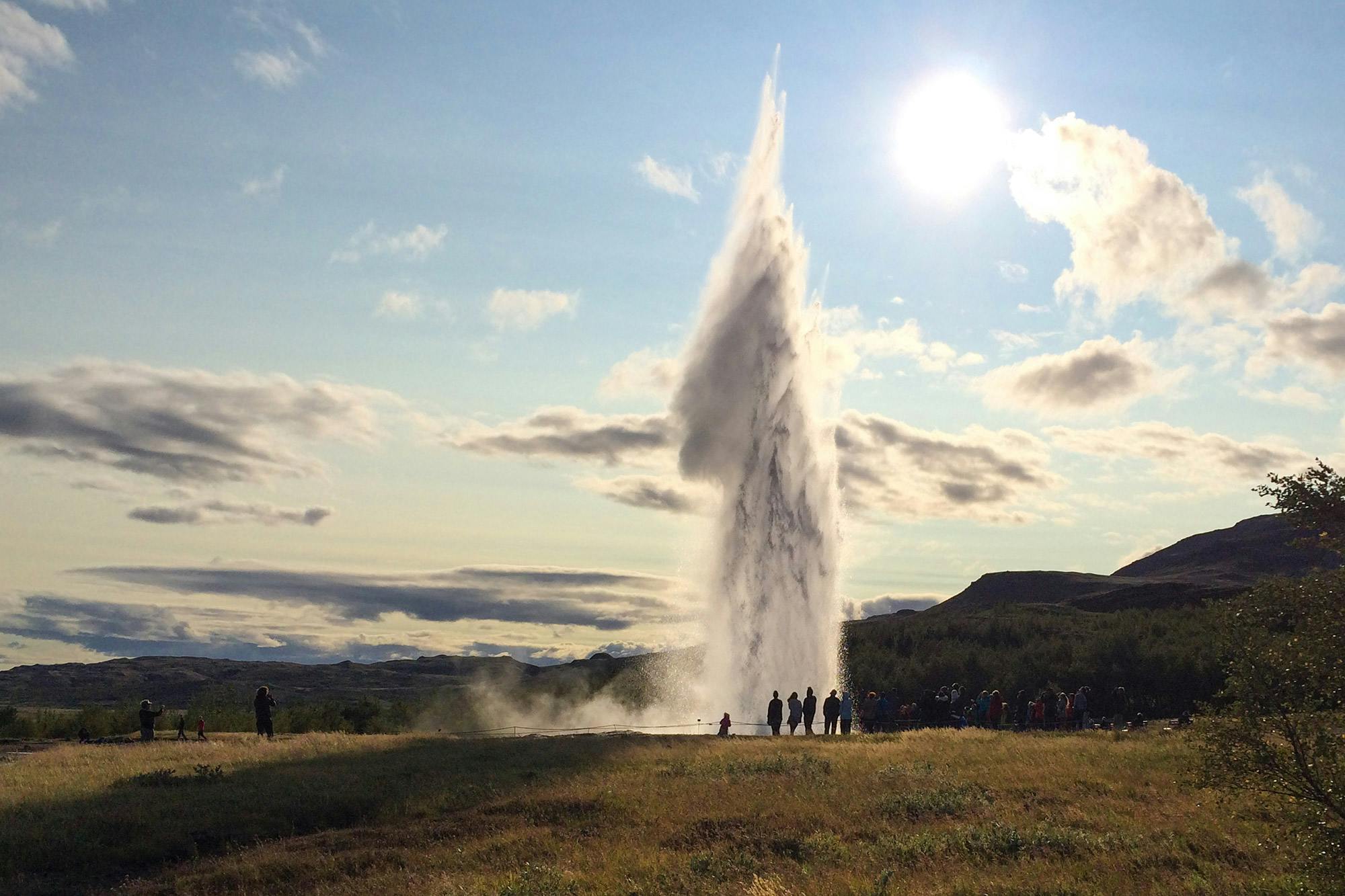 A geyser erupts, shooting water high into the sky, with a group of people watching from a grassy field under a bright sun.