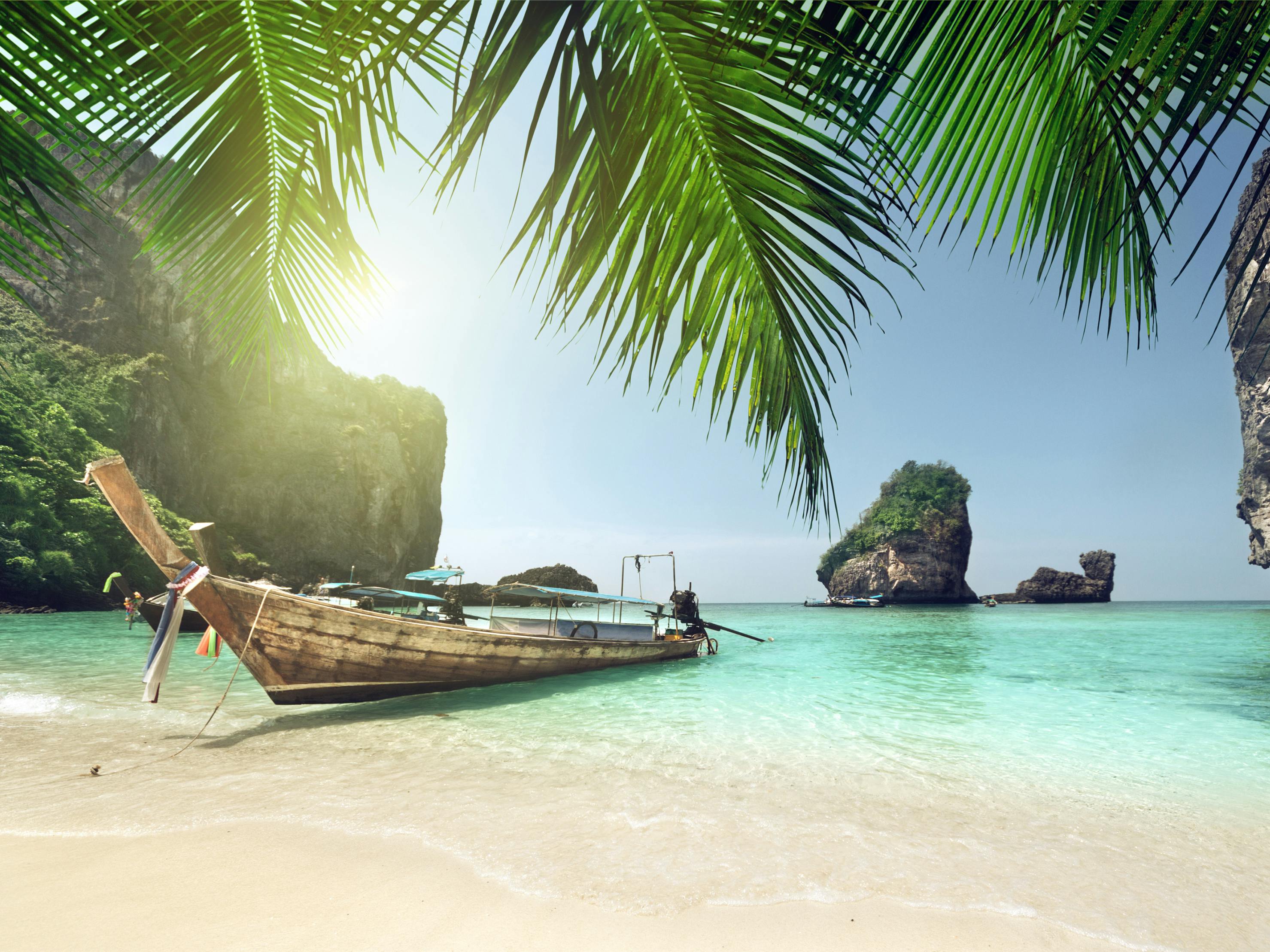 Un bateau en bois est ancré sur une plage tropicale avec une eau claire et turquoise, des îles rocheuses et des feuilles de palmier au-dessus de la tête.