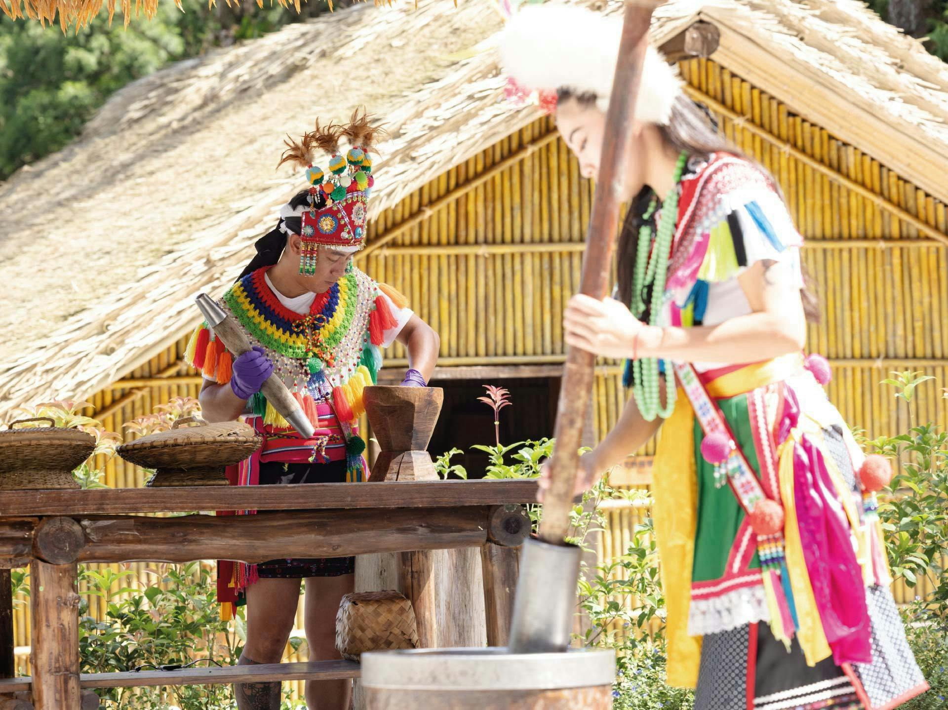 Two individuals in colorful traditional attire working with wooden tools outside a bamboo structure.