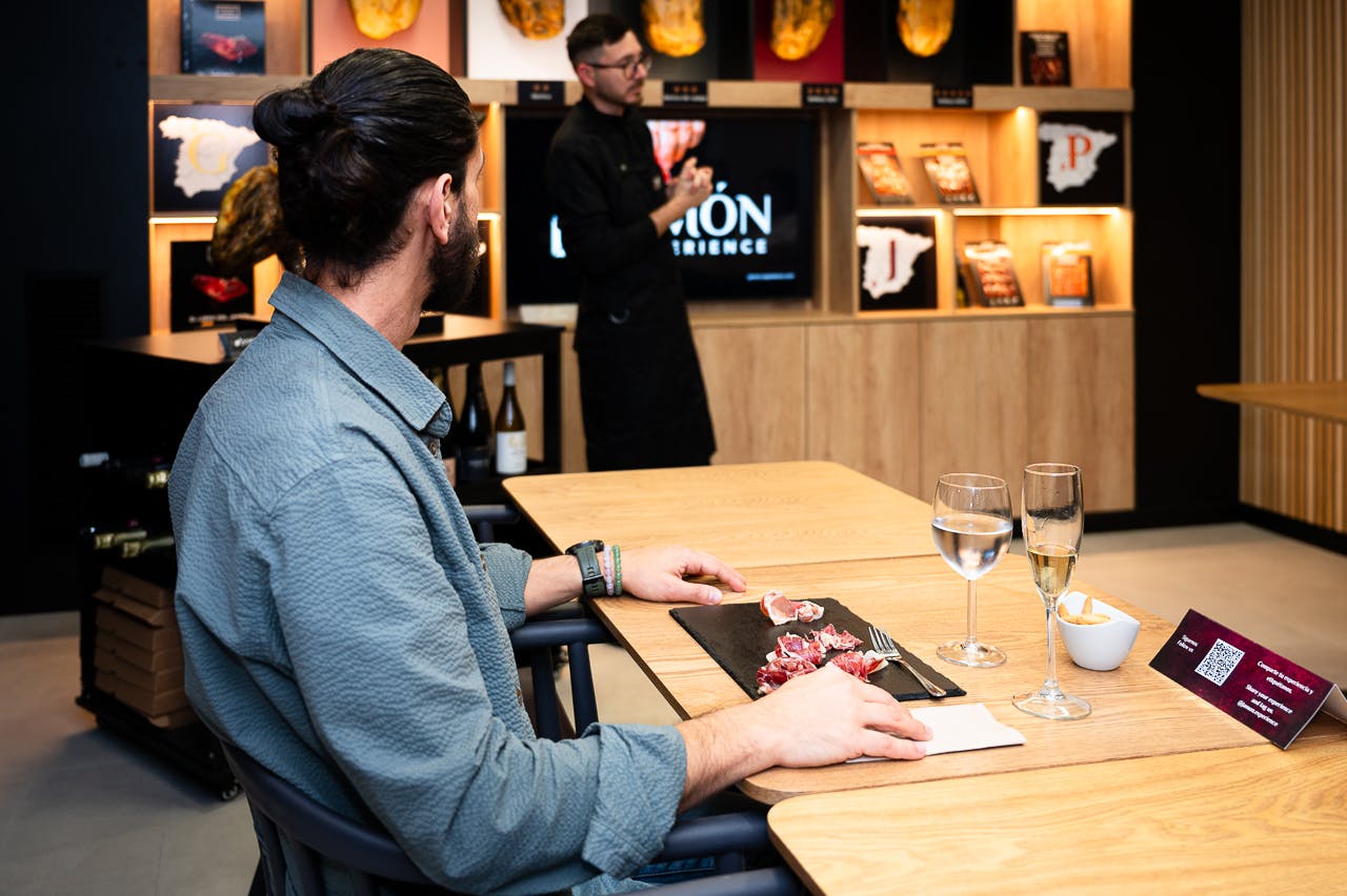 En person, der sidder ved et bord med en tallerken hærdet kød og lytter til en præsentationsvært i en kulinarisk indstilling.