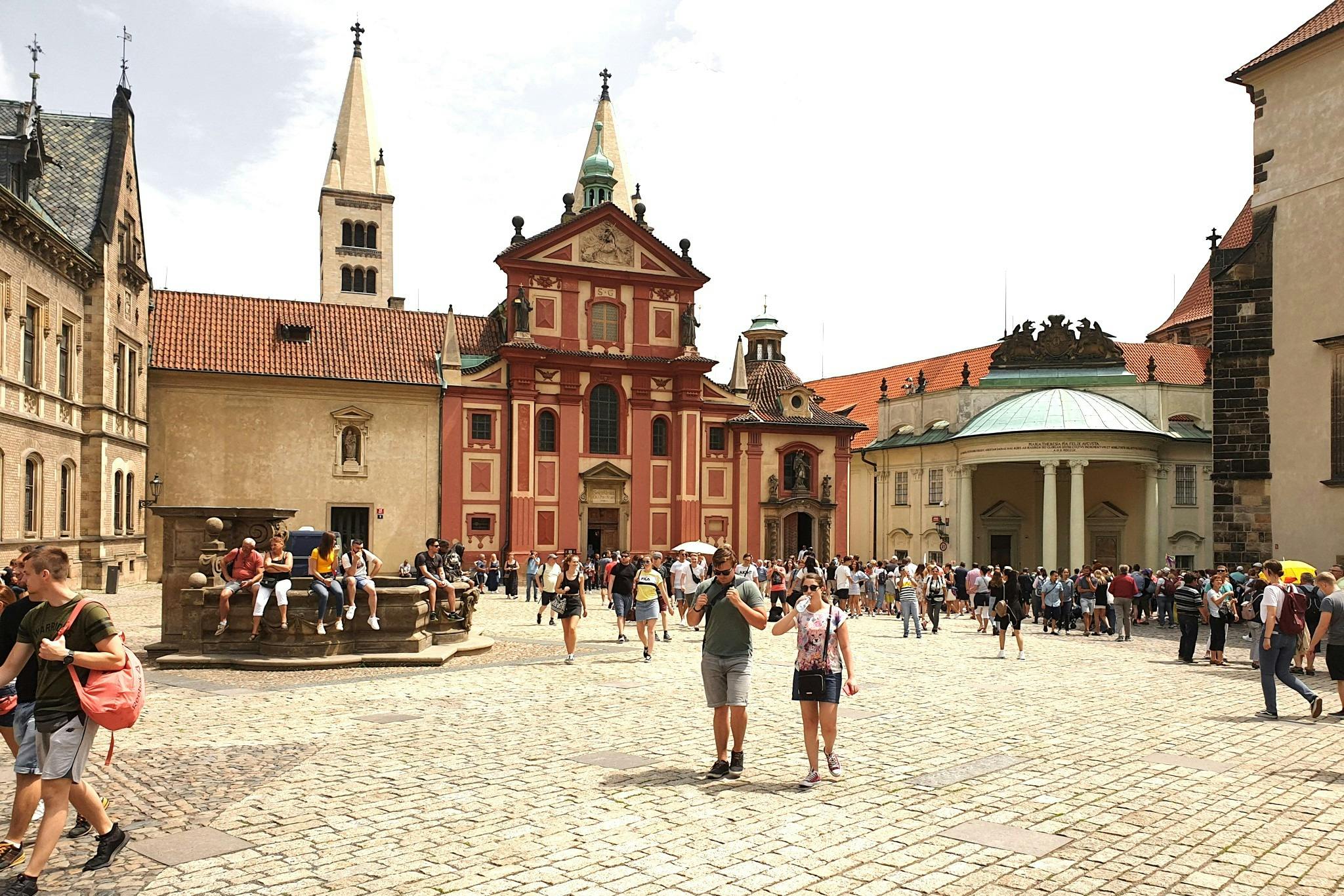 Tourists walk and gather in front of ornate historic buildings with towers and domed roofs in a cobblestone plaza.