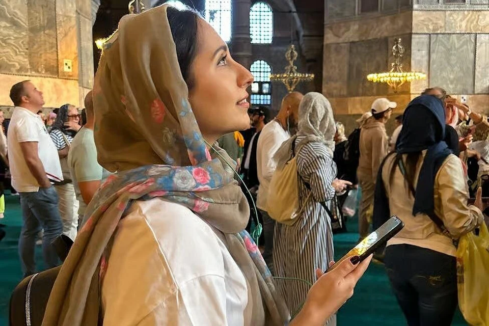 A woman wearing a floral scarf looks upwards while holding a phone in a crowded indoor space with high arched windows.
