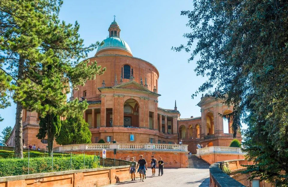 A group of people walk towards a red-brick circular building with a dome, surrounded by trees, under a clear blue sky.