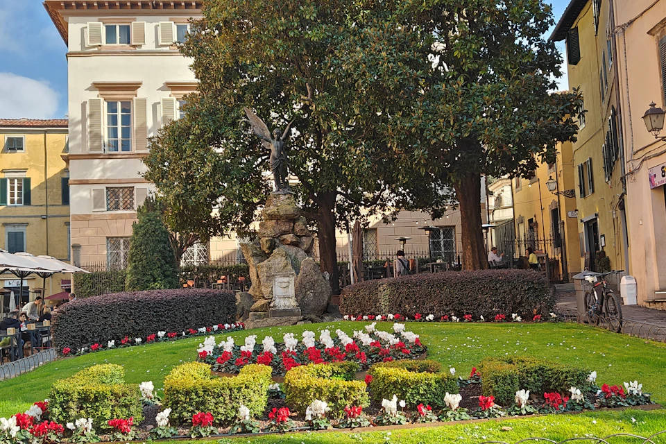 A landscaped garden with colourful flowers, a statue atop rocks, and a background of trees and a building.
