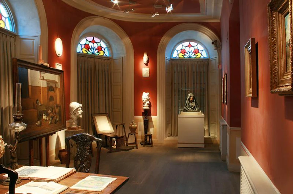 A dimly lit museum room with red walls, stained glass windows, and various sculptures and paintings on display.