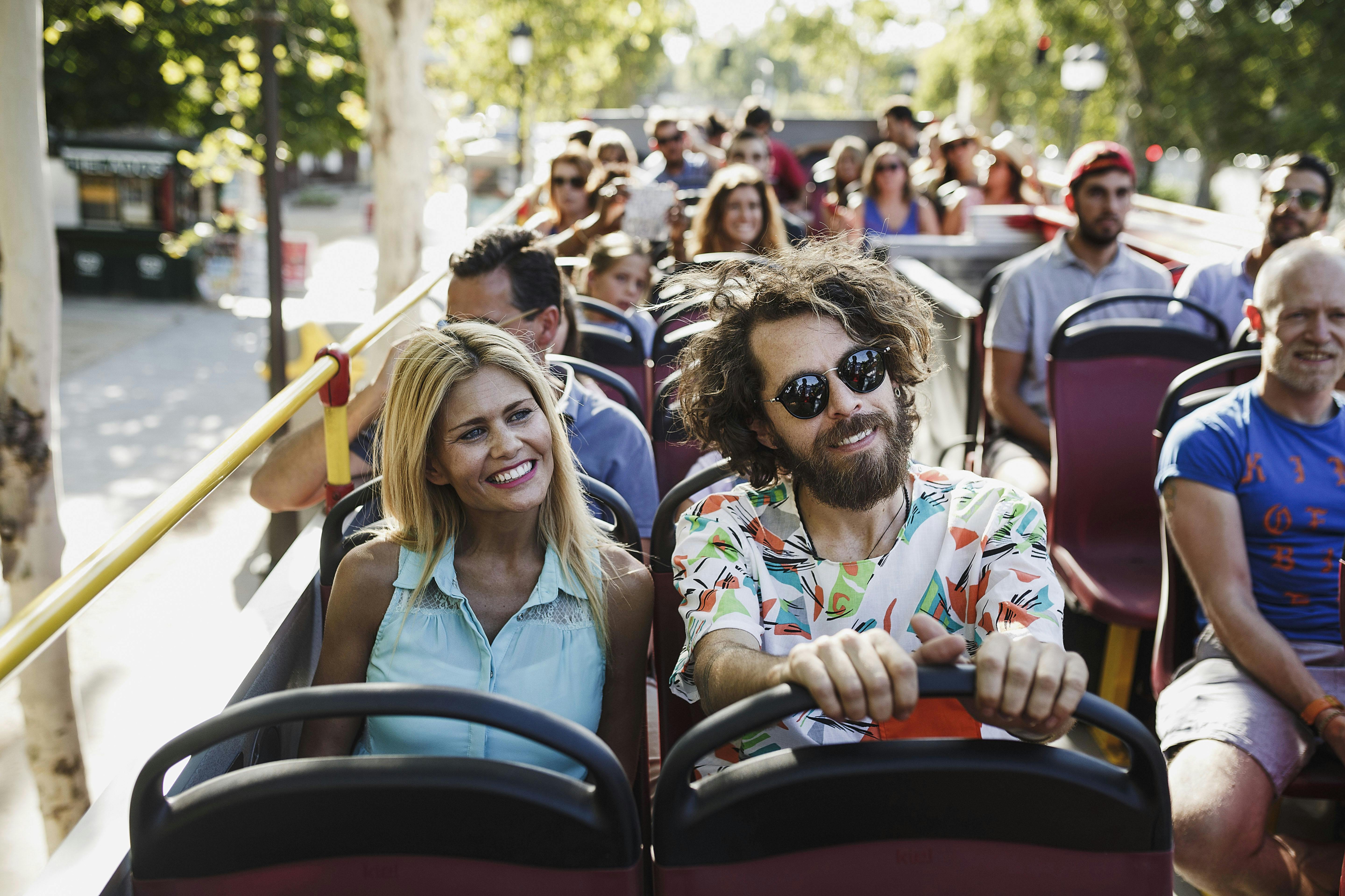 Lidé sedí v turistickém autobuse s otevřenou střechou a užívají si jízdu. Muž vpředu nosí sluneční brýle a barevnou košili.