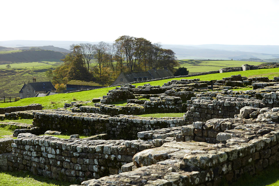 Stone ruins spread across a grassy landscape with distant trees and houses. Rolling hills extend into the background.