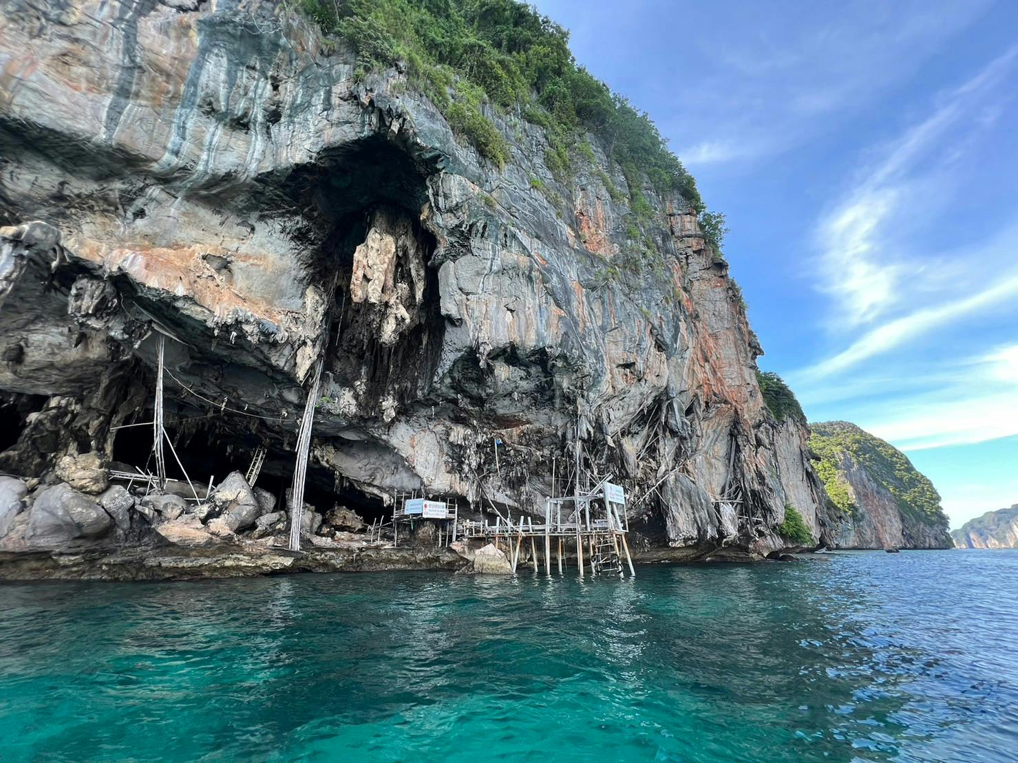 Falaise surplombant une structure en bois près d'une étendue d'eau turquoise, avec une verdure luxuriante au sommet de la falaise sous un ciel bleu.