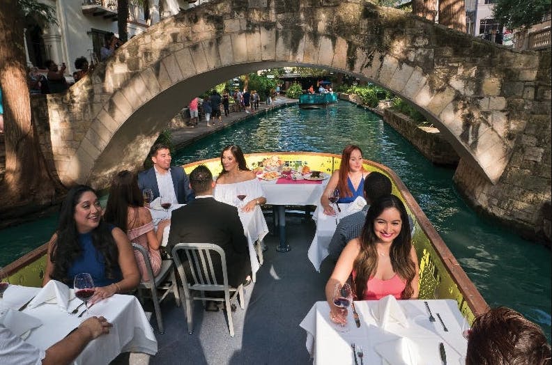 Gent menjant en taules blanques cobertes de llenya en un vaixell de riu sota un pont de pedra, amb un canal i vegetació al fons.