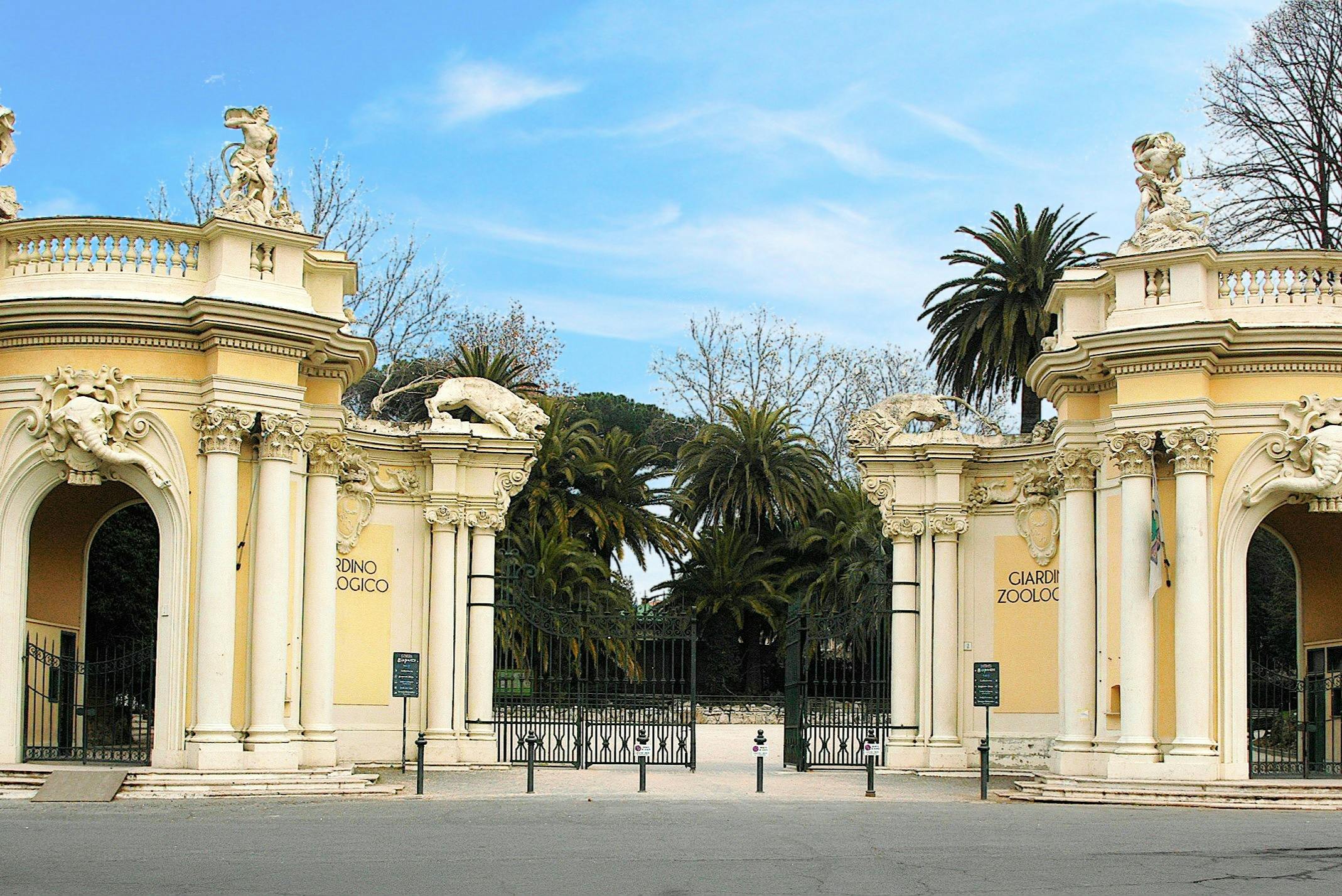 两侧写有“Giardino Zoologico”的华丽入口大门，两侧是蓝天下的雕像和棕榈树。