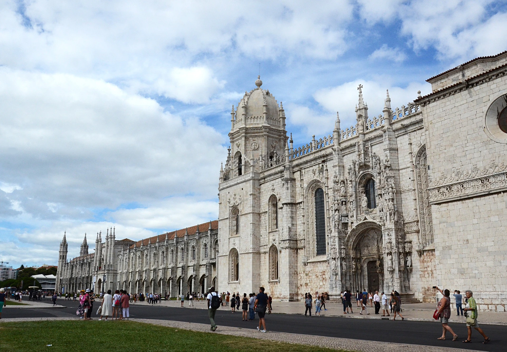 Tourists walking along a majestic, intricately detailed medieval cathedral with arches and a large central dome under a partly cloudy sky.